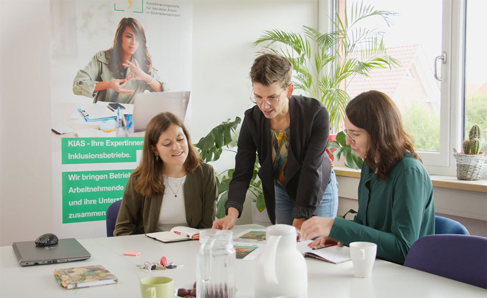 Drei Frauen sitzen und stehen um einen Tisch und besprechen ein Projekt
