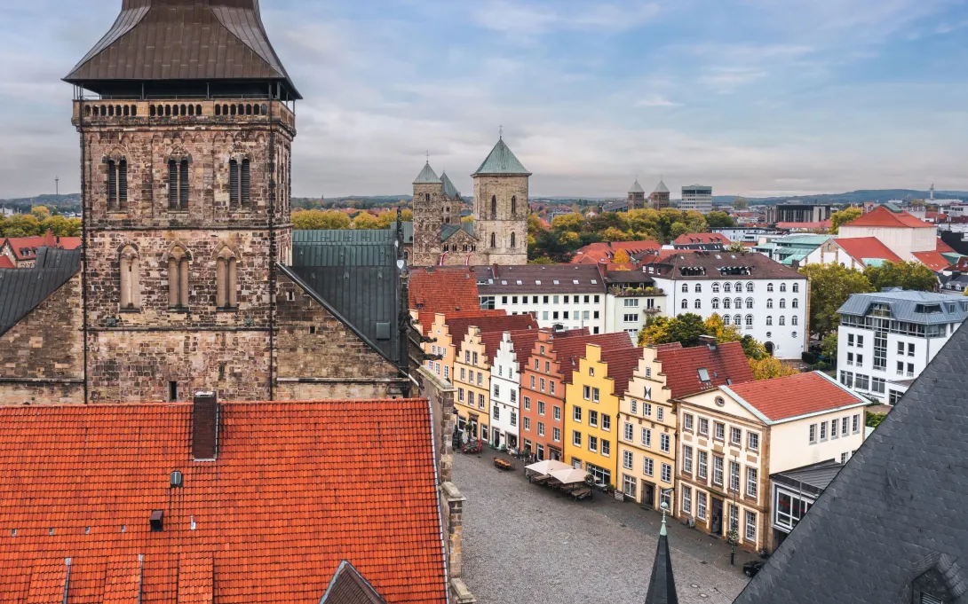 Historische Altstadt mit bunten Giebelhäusern, Kopfsteinpflasterplatz und markanten Kirchtürmen, umgeben von herbstlicher Landschaft.